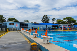 Großer Swimmingpool mit schwimmenden Menschen, umgeben von Bahntrennern, Verkehrskegeln, Stühlen, Sonnenschirmen, einem Gebäude mit Fenstern, einer Flagge, Bäumen und einem bewölkten Himmel.