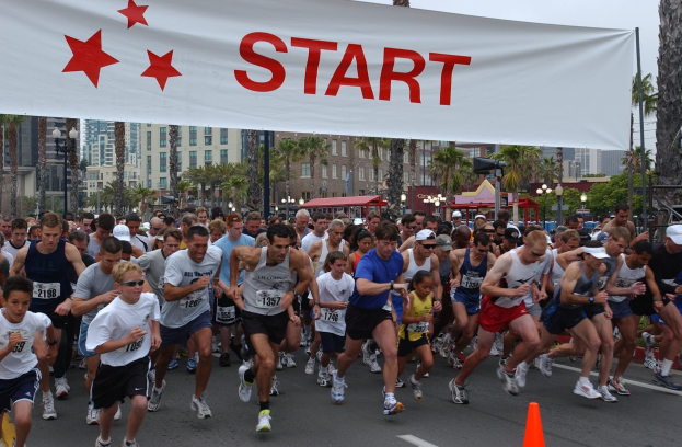 Eine Gruppe von Menschen bei einem Marathon, mit einem Verkehrskegel im Vordergrund und einem Banner mit Text im Hintergrund, umgeben von Bäumen, Laternenmasten, Gebäuden und einem klaren blauen Himmel.