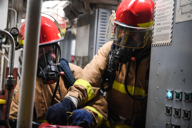 Zwei Feuerwehrleute in Schutzausrüstung und Gasmasken arbeiten an einem Feuerwehrauto, mit einer Tafel mit Text rechts und Metallstangen im Hintergrund.