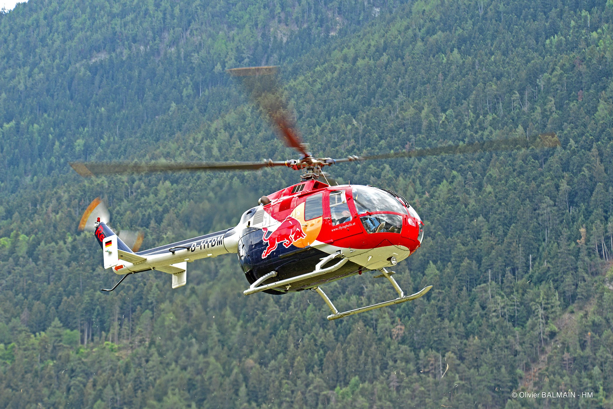 Roter Hubschrauber fliegt über einen grünen Wald mit Hügeln und einem klaren blauen Himmel.