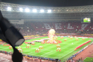 Ein Stadion mit Flutlicht, Eisenstäbe, Zuschauern, stehenden Personen, Werbetafeln, Anzeigetafeln, Überwachungskameras, Fahnen und Dekorationen.
