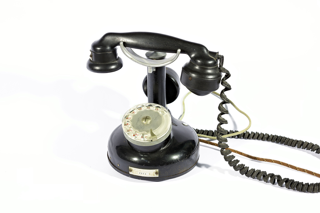 Ein schwarzes Telefon mit Zahlen und Text, der durch die Glasscheibe sichtbar ist, verbunden durch Kabel, vor einem weißen Hintergrund.