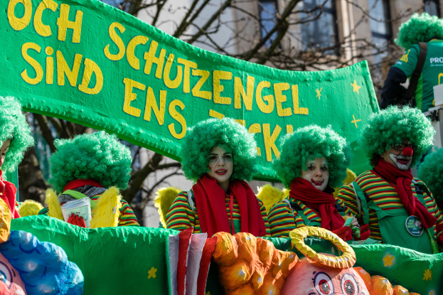 Eine Gruppe von Clowns in bunten Kostümen marschiert durch die Straße und hält ein Banner hoch, mit Bäumen und einem Gebäude im Hintergrund.