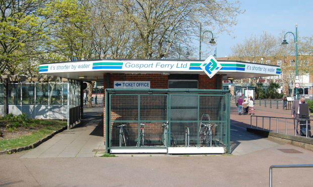 Haltestelle mit 'Gosport Ferry Ltd'-Schild, Metallzaun, Menschen, Fahrrädern, Bäumen, Straßenlaternen, Schildern, einem Gebäude und einem bewölkten Himmel.