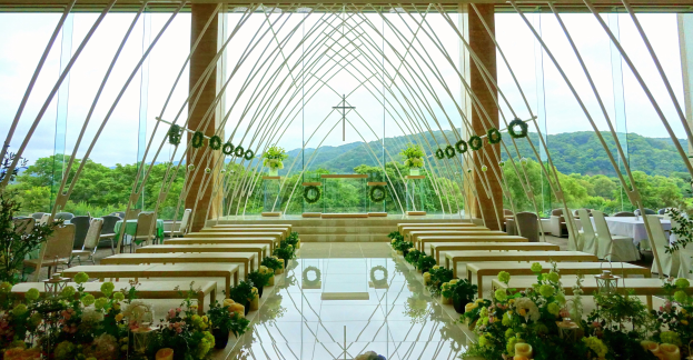 Eine Trauungszeremonie in einem großen Saal mit halbrunder Bestuhlung, geschmückt mit Blumen und anderen Gegenständen, vor einer Kulisse aus Bäumen, Hügeln und einem klaren Himmel.