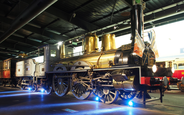 Eine Dampflokomotive in einem Museum ausgestellt, umgeben von anderen Zügen, mit Deckenbeleuchtung und einem Textschild im Hintergrund.