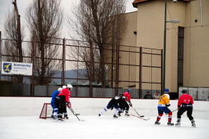 Personen spielen Eishockey auf einem Eisstadion mit Gebäuden, Bäumen und einer Straßenlaterne im Hintergrund.