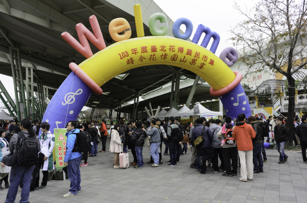 Gruppe von Menschen vor einem Willkommensbogen bei einem Outdoor-Event mit Zelten, Bäumen und Gebäuden unter einem klaren blauen Himmel, einige tragen Taschen.