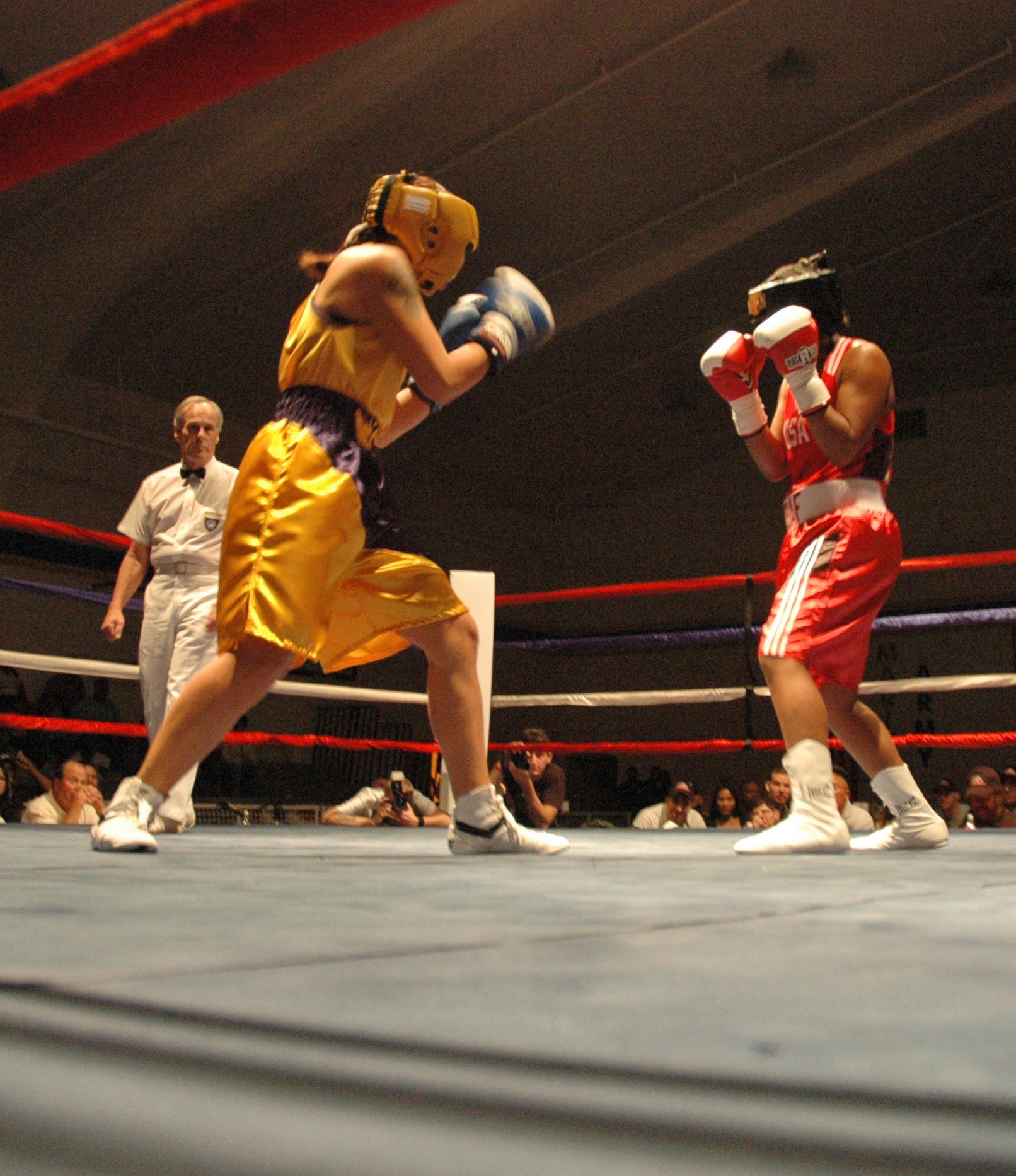 Zwei Boxer im Ring mit einem Schiedsrichter, umgeben von Zuschauern und Fotografen, unter einer sichtbaren Decke.