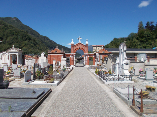Ein Friedhof mit zahlreichen Grabsteinen und Blumen im Vordergrund, ein Tor im Hintergrund, umgeben von Gebäuden, Bäumen, Hügeln und einem bewölkten Himmel.