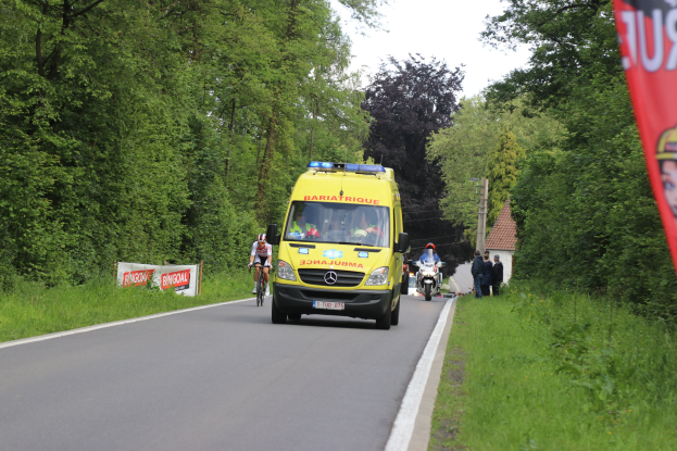 Ambulanz fährt auf einer Straße mit Fahrradfahrern nebenher, Gras und Bäume auf beiden Seiten, Häuser und Strommasten im Hintergrund unter einem klaren blauen Himmel.