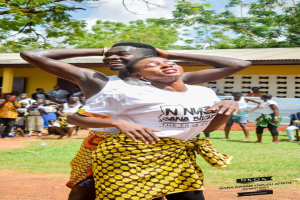 Ein Mann in einem weißen und gelben Kleid tanzt vor einer Menge, mit einem Haus, Bäumen und einem bewölkten Himmel im Hintergrund.