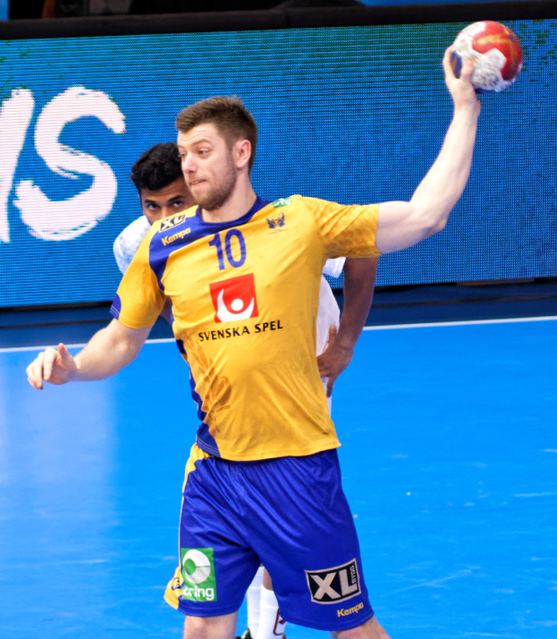 Ein Mann in einem gelben T-Shirt und blauen Shorts hält einen Handball auf einem Feld, im Hintergrund ist ein Bildschirm zu sehen, auf dem "Svenský Spel Handball World Cup 2019" steht.