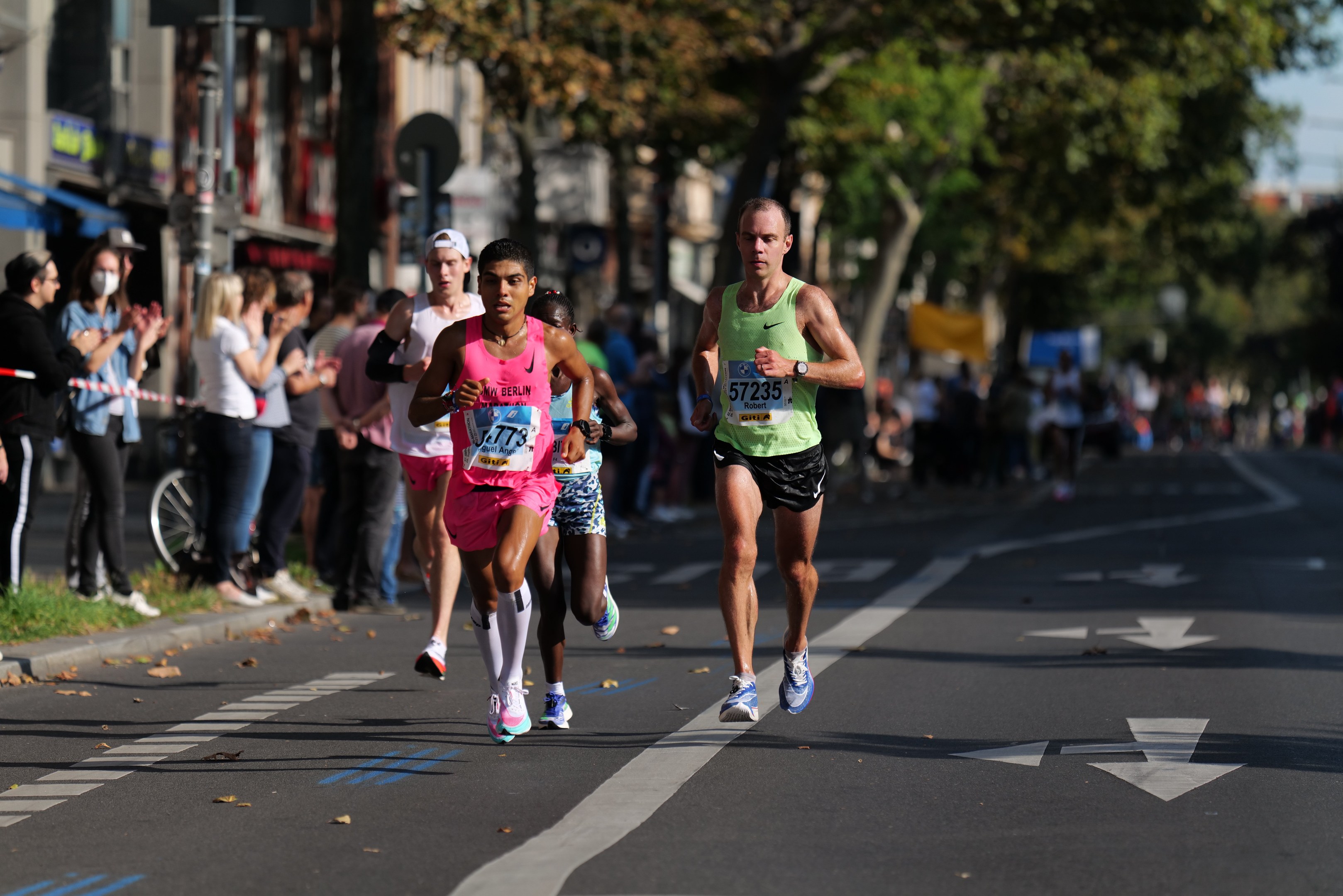 Gruppe von Menschen, die bei einem Marathon auf einer Stadtstraße laufen, mit Zuschauern auf der linken Seite, unscharfen städtischen Hintergrund mit Bäumen, Gebäuden und einem Fahrrad.