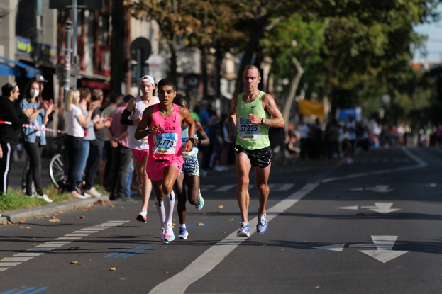 Gruppe von Menschen, die bei einem Marathon auf einer Stadtstraße laufen, mit Zuschauern auf der linken Seite, unscharfen städtischen Hintergrund mit Bäumen, Gebäuden und einem Fahrrad.