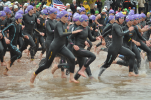 Menschen in Badeanzügen durch flaches Wasser rennen, während andere im Hintergrund stehen und eine Fahne zu sehen ist.