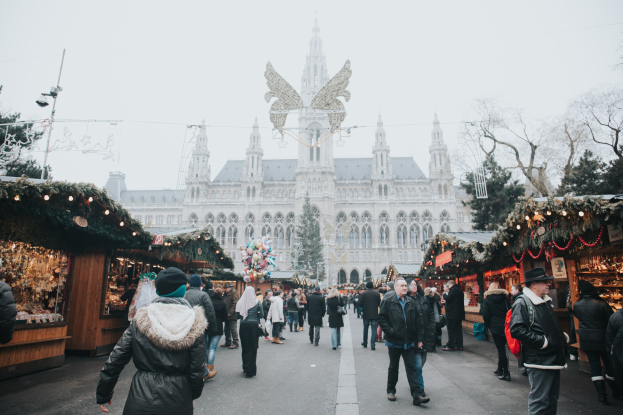Ein geschäftiges Weihnachtsmarkt in Wien, Österreich, mit Menschen, die herumlaufen, Ständen, die mit Lichtern und festlichen Artikeln dekoriert sind, einem Gebäude mit Fenstern, Bäumen und einem klaren blauen Himmel im Hintergrund.