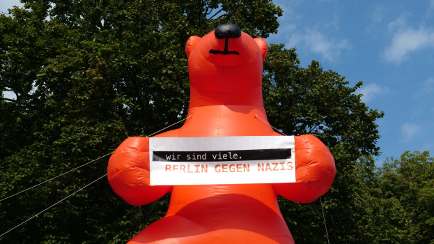 Aufblasbarer Bär mit einem Schild "Berlin gegen Nazis" vor Bäumen und einem bewölkten Himmel.