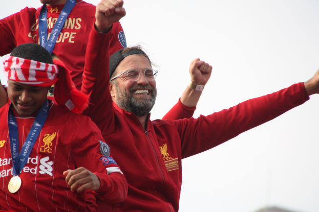 Liverpool-Trainer Jürgen Klopp feiert mit Spielern und Staff nach dem Gewinn der UEFA Champions League, trägt eine rote Jacke und ist von zwei medaillentragenden Personen umgeben, während der Himmel sichtbar ist.
