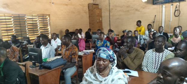 Eine Gruppe von Menschen sitzt an Tischen in einem Klassenzimmer mit Monitoren, Taschen, Büchern, Papieren, mit einem Fenster auf der linken Seite, Kartons und einem Fernseher im Hintergrund und Text unten, der eine Schulung zur Verwendung von Technologie anzeigt.