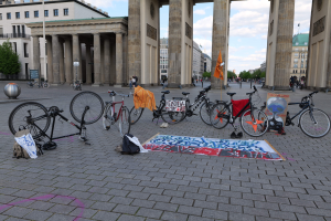 Eine Gruppe von Fahrr├Ądern, die vor dem Brandenburger Tor mit Taschen und einer Fahne in der N├Ąhe geparkt sind, vor einem Hintergrund aus Geb├Ąuden, B├Ąumen und einem klaren Himmel.
