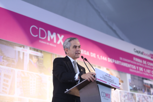 Ein Mann im Anzug und Krawatte steht an einem Podium mit zwei Mikrofonen und einer Tafel vor ihm, mit einem Banner mit Bildern und Text dahinter.