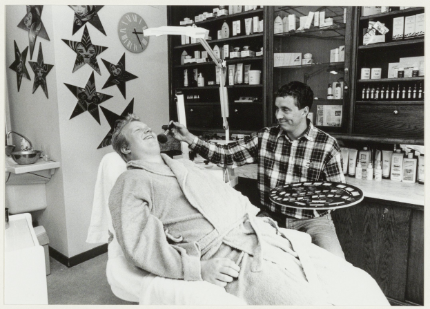 Ein Mann sitzt lachend in einem Zahnarztstuhl, während ein anderer Mann neben ihm steht und ein Tablett hält, mit einem Waschbecken, Sternen, einer Uhr und Regalen mit Flaschen im Hintergrund.