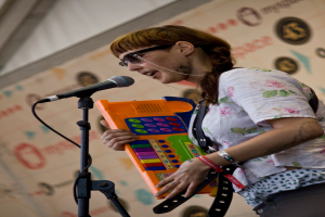 Eine Frau in einem weißen Hemd und Sonnenbrille singt in ein Mikrofon und spielt Klavier, während ein Plakat an der Wand hinter ihr zu sehen ist.