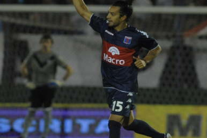 Ein Fussballspieler feiert nach einem Tor auf dem Boden, mit jubelnden Zuschauern und einer Person im Hintergrund, alles vor einem leicht unscharfen Hintergrund.