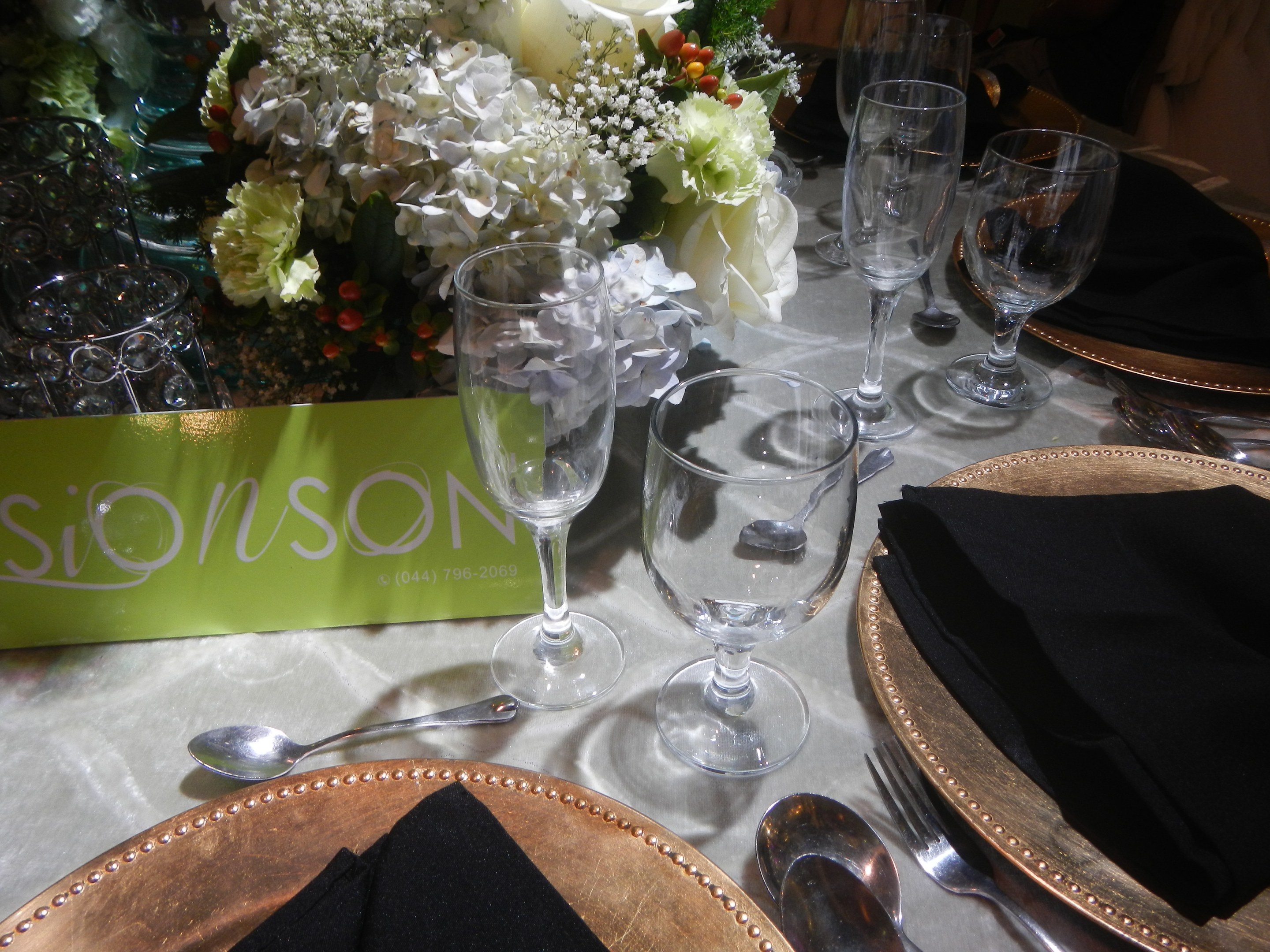 Formaler Esstisch mit goldenen Untersetzern, schwarzen Servietten, Besteck, Gläsern und einem Blumenstrauß, mit einer Texttafel.