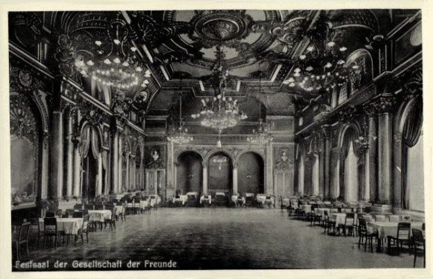 Ein altes Schwarz-Weiß-Foto eines großen Raums mit Tischen, Stühlen, Säulen, Bögen und Kronleuchtern an der Decke, mit Text unten, der "Fest der Gesellschaft der Freunde, Berlin" lautet.