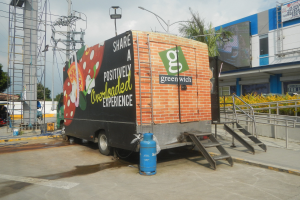 Ein großer Lastwagen steht vor einem Gebäude mit einer Tafel, auf der "Teilen Sie eine positiv überladene Erfahrung" steht, mit Stufen, einem Zylinder auf dem Boden, Geländern, Strommasten, Bäumen und einem bewölkten Himmel.