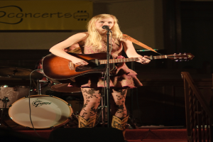 Eine Frau, die auf der Bühne Gitarre spielt und singt, mit einer Band im Hintergrund und einem Bildschirm an der Wand hinter ihr.