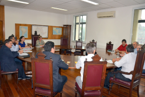 Eine Gruppe von Menschen, die an einem Tisch mit Papieren und Gläsern sitzt, ein Klimagerät, eine Anschlagtafel und Karten an der Wand im Hintergrund.