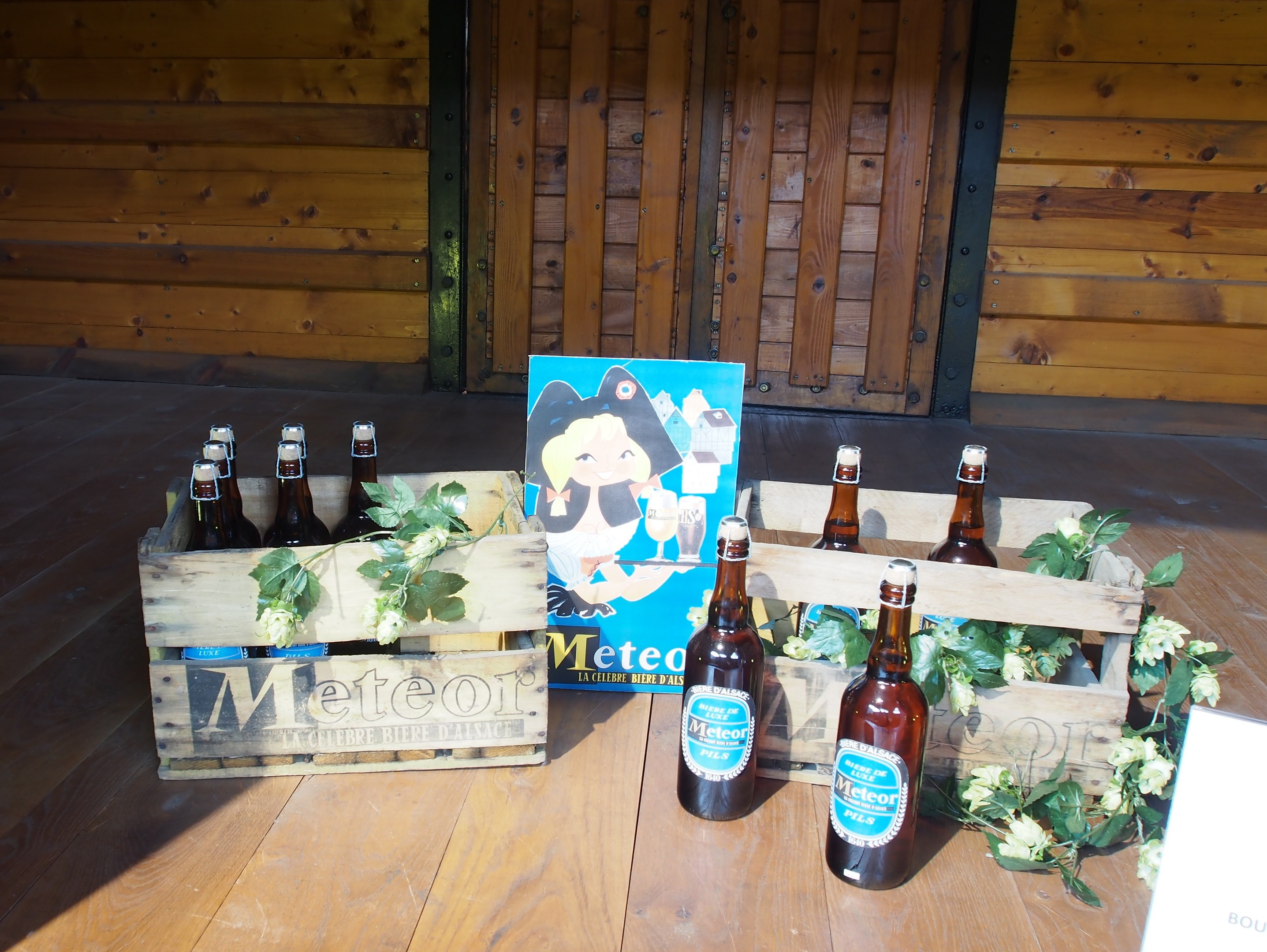 Ein hölzerner Kasten voller Bierflaschen auf einem hölzernen Boden, umgeben von Blättern und Blumen, mit einer hölzernen Wand und Tür im Hintergrund.