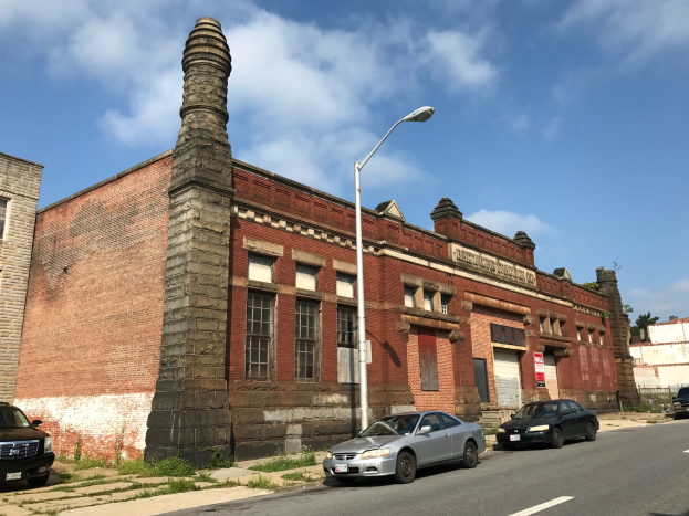 Alte Backstein-Feuerwache-Gebäude mit seitlichem Schornstein, umgeben von Straßenmasten, Laternen, Fahrzeugen, Schildern, Bäumen und einem bewölkten Himmel.