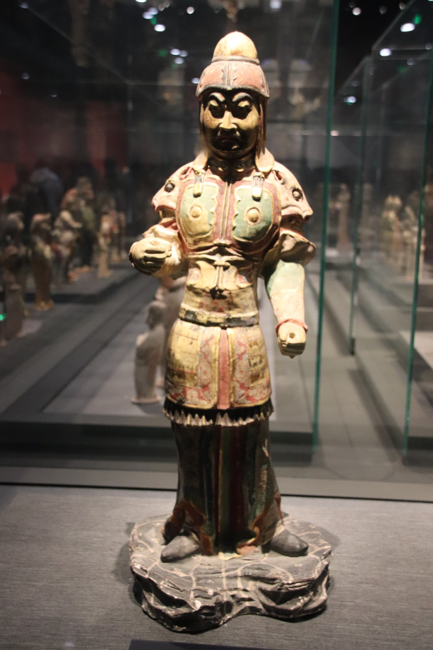 Terrakotta-Krieger-Statue in einer Glasvitrine mit detaillierter Rüstung, ausgestreckten Armen und einem friedlichen Gesicht, beleuchtet von Hintergrundlichtern.