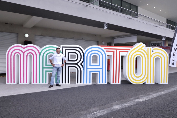 Ein Mann steht am Start eines Marathons vor einem großen "Marathon"-Schild, mit einer Fahne rechts und einem Gebäude mit Lampen, Geländern und Fensterläden im Hintergrund.