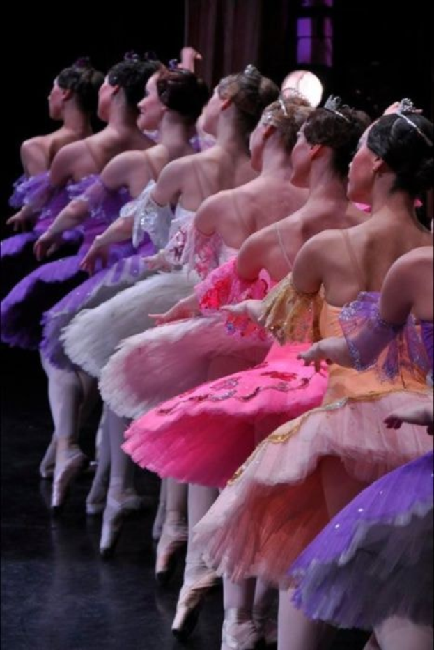 Eine Gruppe von Balletttänzerinnen in rosafarbenen Tutus führt synchronisierte Bewegungen auf einer Bühne auf, beleuchtet von einem Scheinwerfer im Hintergrund.