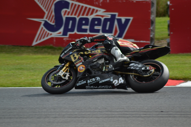 Eine Person in Helm und Schuhen fährt ein Motorrad auf einer Rennstrecke, im Hintergrund ist Gras und eine Texttafel zu sehen.