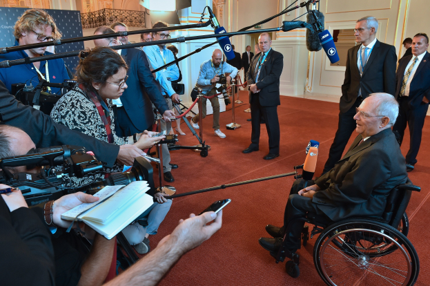 Ein Mann im Anzug und Krawatte sitzt im Rollstuhl, umgeben von Reportern mit Kameras und Mikrofonen, mit einem Banner und einer Tür im Hintergrund.