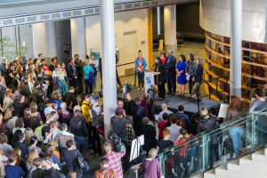 Eine Gruppe von Menschen steht auf einer Treppe, die zu einem Podium mit einem Mikrofon führt, und schaut in eine Menge mit Kameras auf einem Event mit Säulen, einer Tür und einer Topfpflanze.
