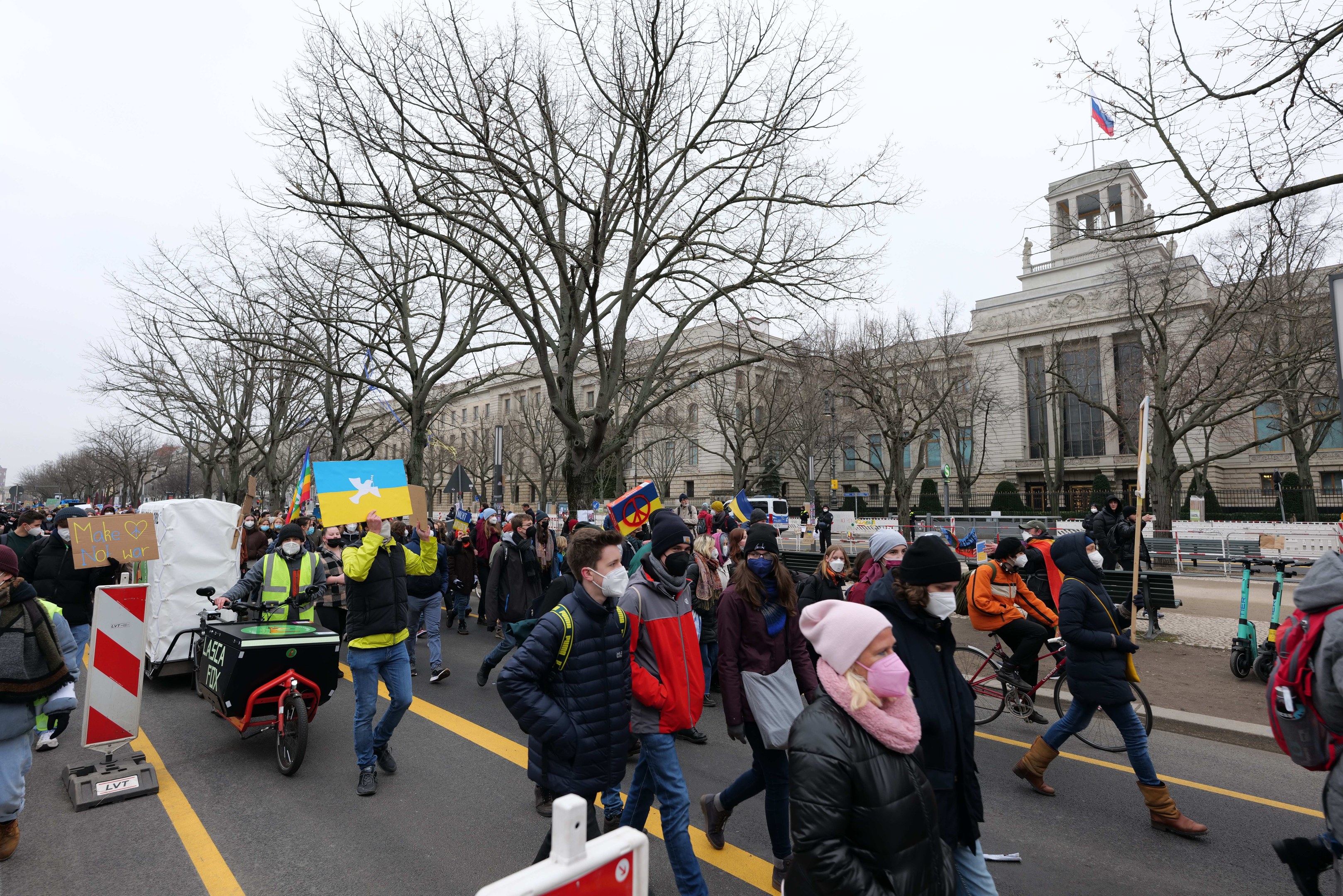 Eine große Gruppe von Menschen marschiert auf einer Demonstration eine Straße in Washington, D.C. entlang, einige halten Schilder und andere fahren Fahrräder, mit Bäumen und einem Gebäude im Hintergrund unter einem klaren blauen Himmel.