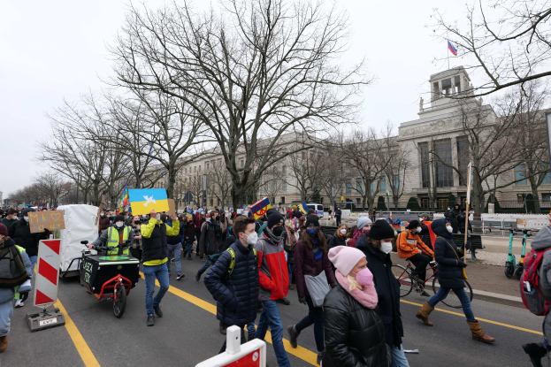 Eine große Gruppe von Menschen marschiert auf einer Demonstration eine Straße in Washington, D.C. entlang, einige halten Schilder und andere fahren Fahrräder, mit Bäumen und einem Gebäude im Hintergrund unter einem klaren blauen Himmel.