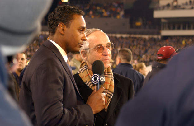 Ein Mann im Anzug und Krawatte spricht in ein Mikrofon und spricht mit einem Reporter, im Hintergrund ist eine unscharfe Menge in einem Stadion zu sehen.