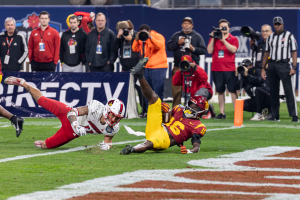 Ein Football-Spieler taucht in die Endzone ein, um einen Touchdown zu erzielen, mit einer Gruppe von Menschen am Boden, einige halten Kameras, und ein Banner mit Text im Vordergrund und Geländern und Pfosten im Hintergrund.