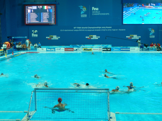 Wasserballspieler bei den FINA-Weltmeisterschaften 2016 in Kazan im Pool gegeneinander antretend, mit Zuschauern auf der linken Seite und einer Anzeigetafel mit Event-Branding im Hintergrund.