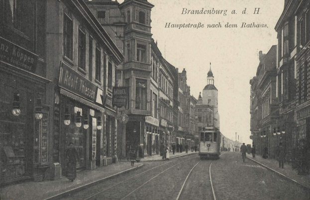 Ein altes Schwarz-Weiß-Foto einer Straßenkreuzung in Brandenburg, Deutschland, mit einer Straßenbahn auf den Schienen, Menschen auf dem Gehweg, Gebäuden mit Fenstern, Schildern und Straßenlaternen unter einem bewölkten Himmel.