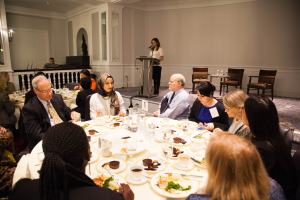 Gruppe von Menschen an einem Esstisch mit Essen und Besteck während einer Besprechung, mit einer Frau, die im Hintergrund an einem Podium spricht.