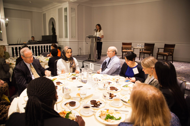 Gruppe von Menschen an einem Esstisch mit Essen und Besteck während einer Besprechung, mit einer Frau, die im Hintergrund an einem Podium spricht.